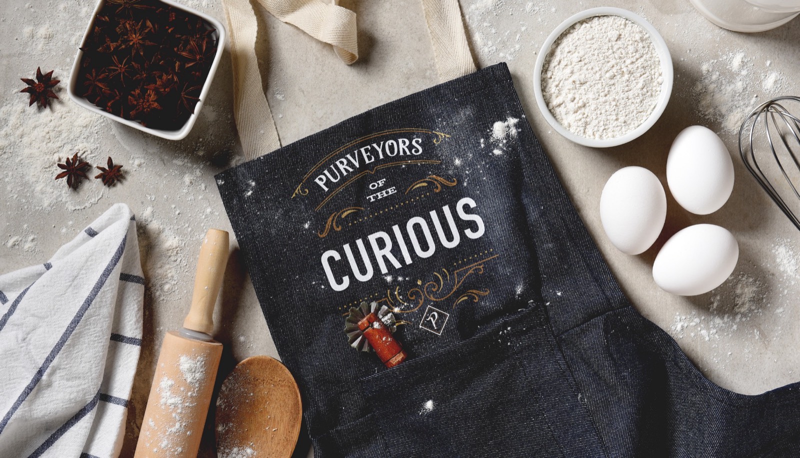 Denim staff apron printed with Purveyors of the Curious, surrounded by baking ingredients