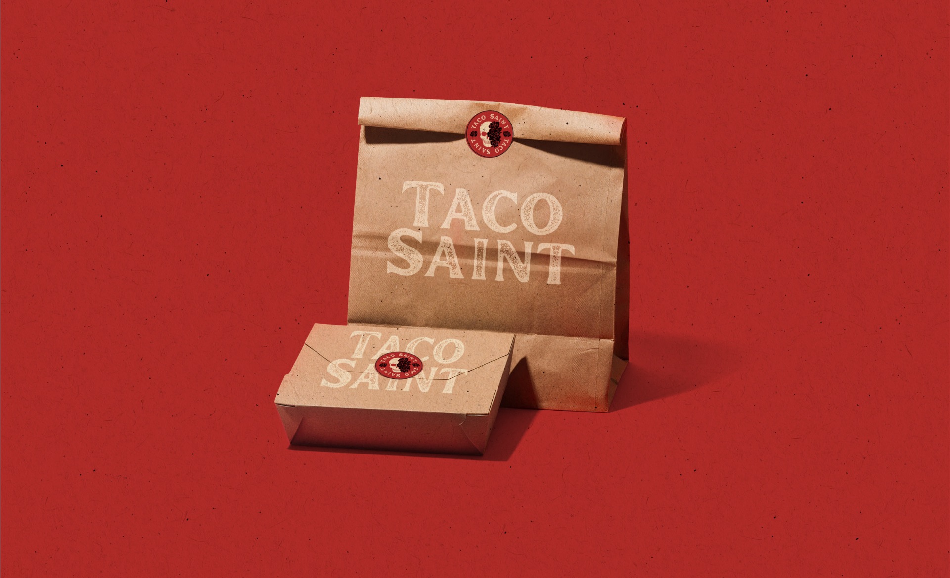 Taco Saint kraft takeout bag and box with the skull-and-roses seal on a red background