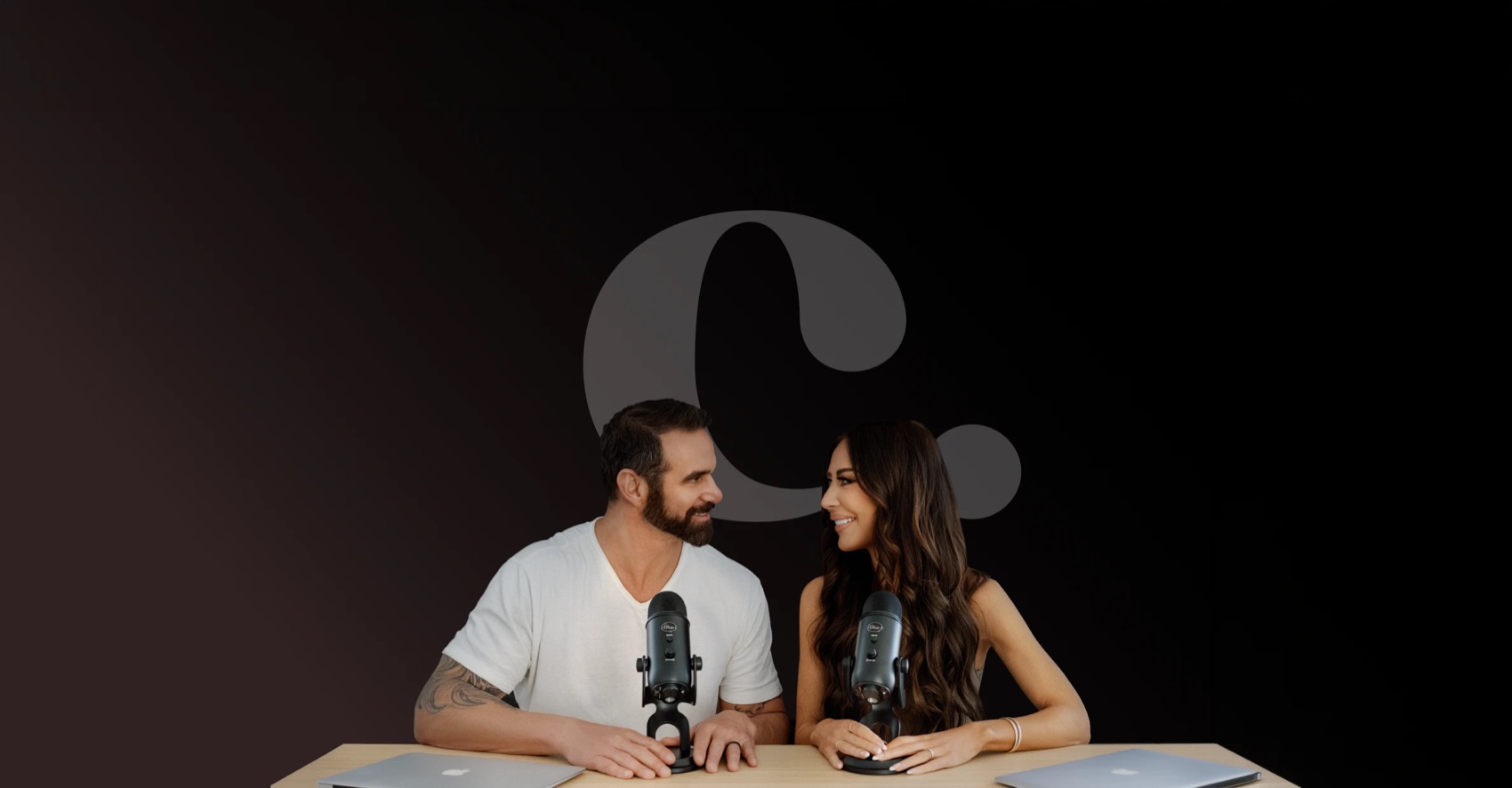 Marcus and Amber Capone at podcast microphones in front of the c. mark